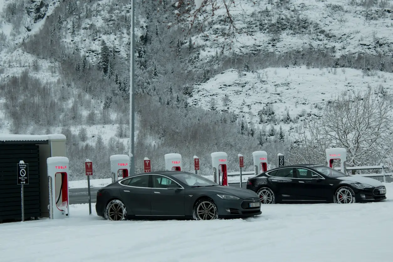 Two Teslas charging at snowy Superchargers, mountains in background.