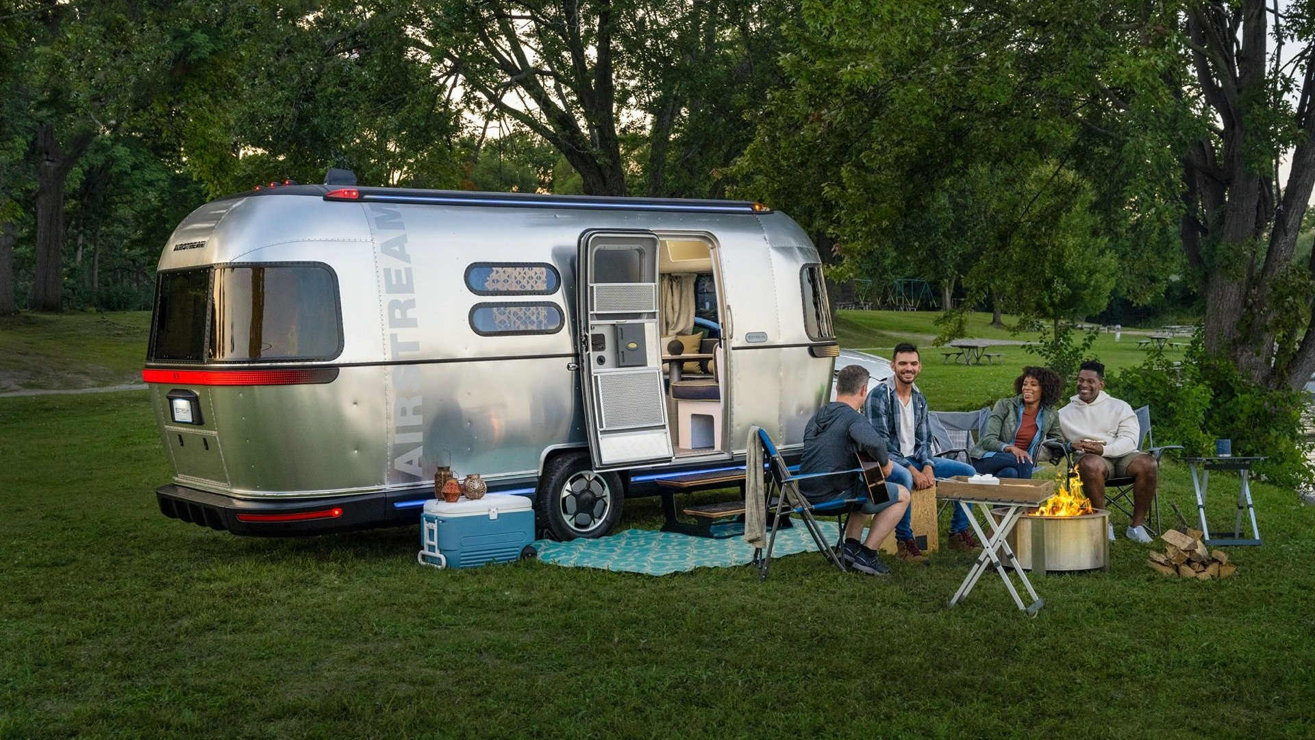 Friends camping by Airstream trailer, roasting marshmallows around fire pit. 